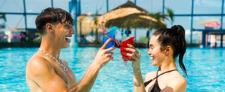 Two people holding colorful drinks, smiling and lounging in a refreshing pool.