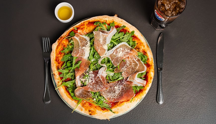 Italian pizza with cured ham, arugula and parmesan served with olive oil and coke on a black tray.