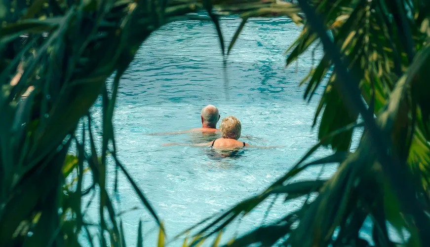 Photo of an elderly couple relaxing in a swimming pool, surrounded by palm leaves, viewed from hiding through dense tropical vegetation