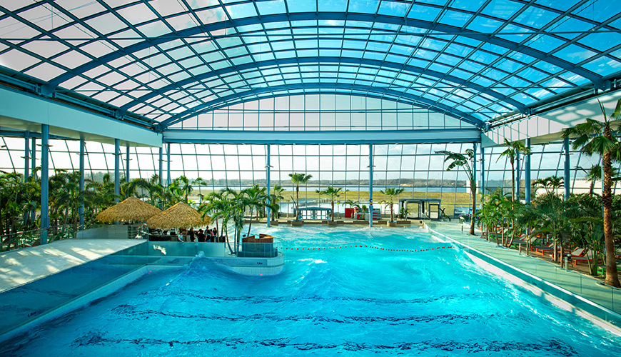 A spacious indoor pool with a roof overhead, surrounded by palms and bright lighting.