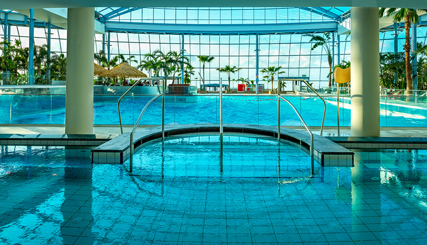 A spacious indoor pool with a roof overhead, surrounded by palms and bright lighting.