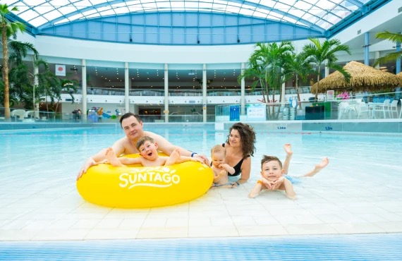 A family of four enjoys playing on a yellow raft in a large pool, with palm trees visible in the background and Jamango Suntago logo.