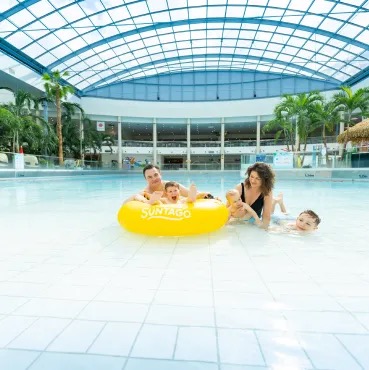 A family of four enjoys playing on a yellow raft in a large pool, with palm trees visible in the background and Jamango Suntago logo.