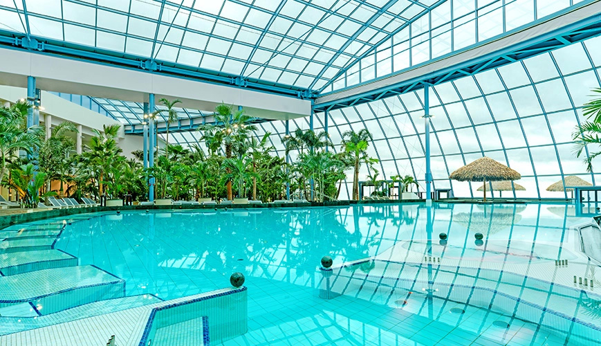 Bright indoor pool featuring a glass roof, surrounded by palm trees and in-water beds for relaxation.