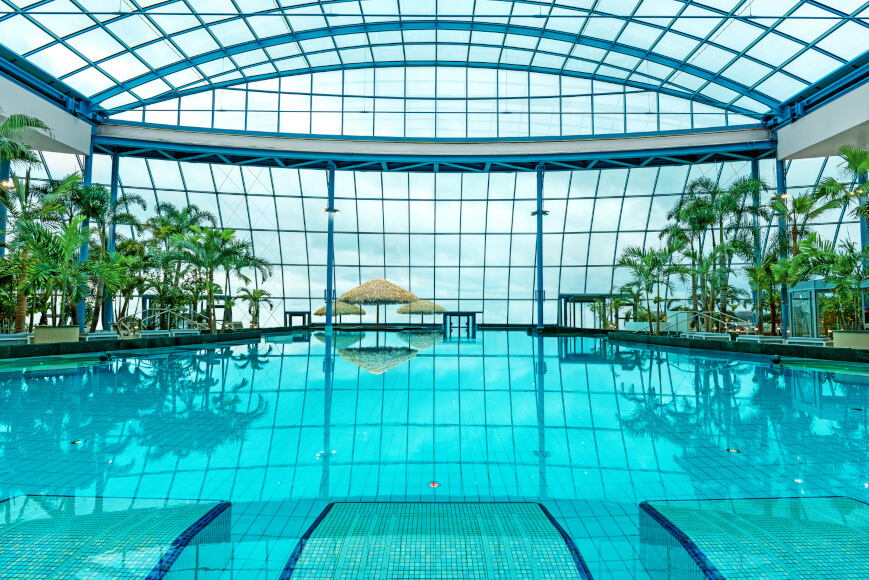 Interior of a tropical swimming pool with clear blue water, surrounded by palm trees. The facility is housed under a large glass dome, with thatched parasols visible in the background.
