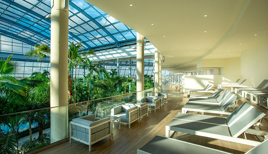 Modern relaxation area with loungers and armchairs lined up along a glass railing. The space is bright, overlooking exotic plants and a pool under a glass roof.