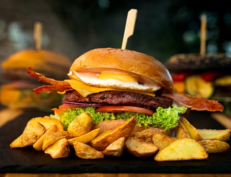 A hamburger with crispy bacon, fresh lettuce, and ripe tomato, served on a stylish black slate plate with grilled potatoes on the side.