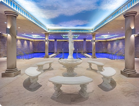 An indoor area with antic-styled sculpture surrounded by benches, leading to mineral pools, under a sky-themed ceiling.
