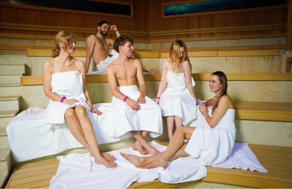 A group of five friends relaxes in a sauna, sitting on wooden benches, with the Saunaria Suntago logo in the background.