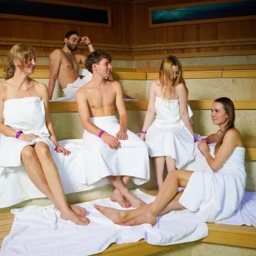 A group of five friends relaxes in a sauna, sitting on wooden benchesr, with the Saunaria Suntago logo in the background.